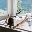 A person relaxing in a bathtub with champagne, in a bright bathroom with scenic views, aligned with Walker Plumbing services.