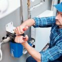 Technician repairing plumbing equipment indoors in a blue plaid shirt and cap for Walker Plumbing, Heating & Air.