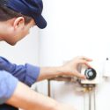 HVAC technician in uniform servicing a wall-mounted thermostat, with visible heating system pipes and tools nearby.