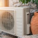 Air conditioning unit by foliage and clay pot on a textured stone wall, representing Walker Plumbing, Heating & Air.